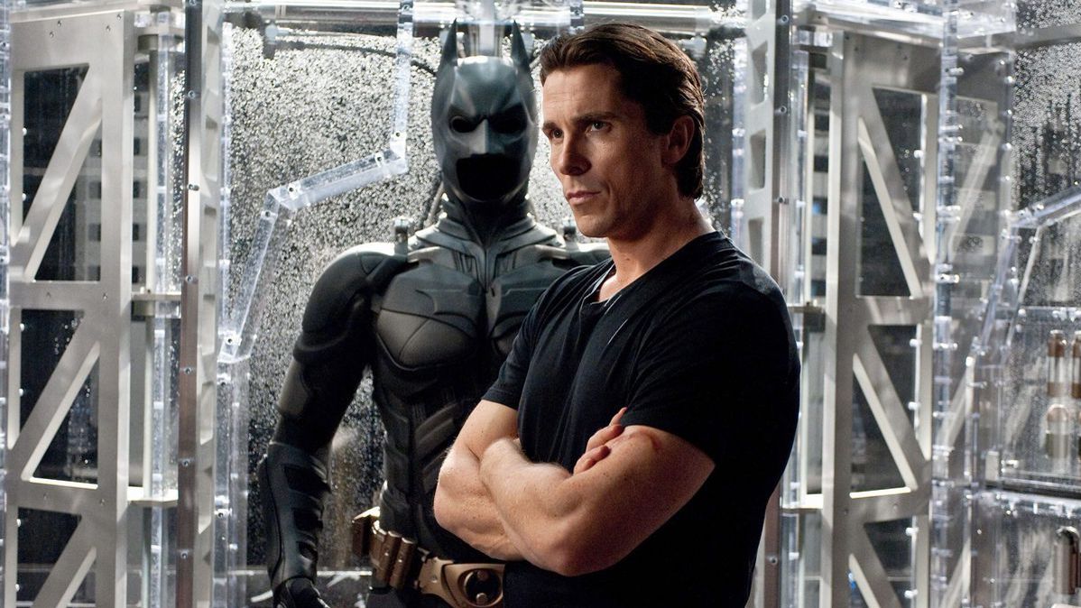 Bruce Wayne crosses his arms as he stands in front of a batsuit in The Dark Knight Rises