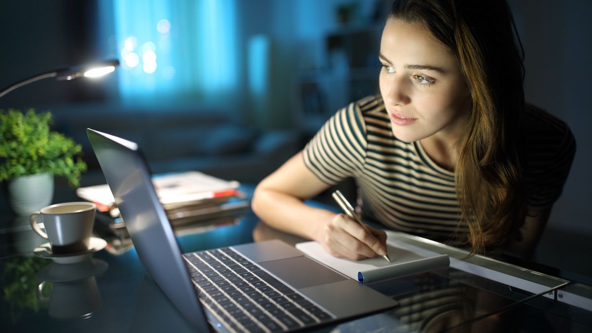 A woman sitting at a table, looking at her laptop on the table and writing something down on paper, while in a dark room